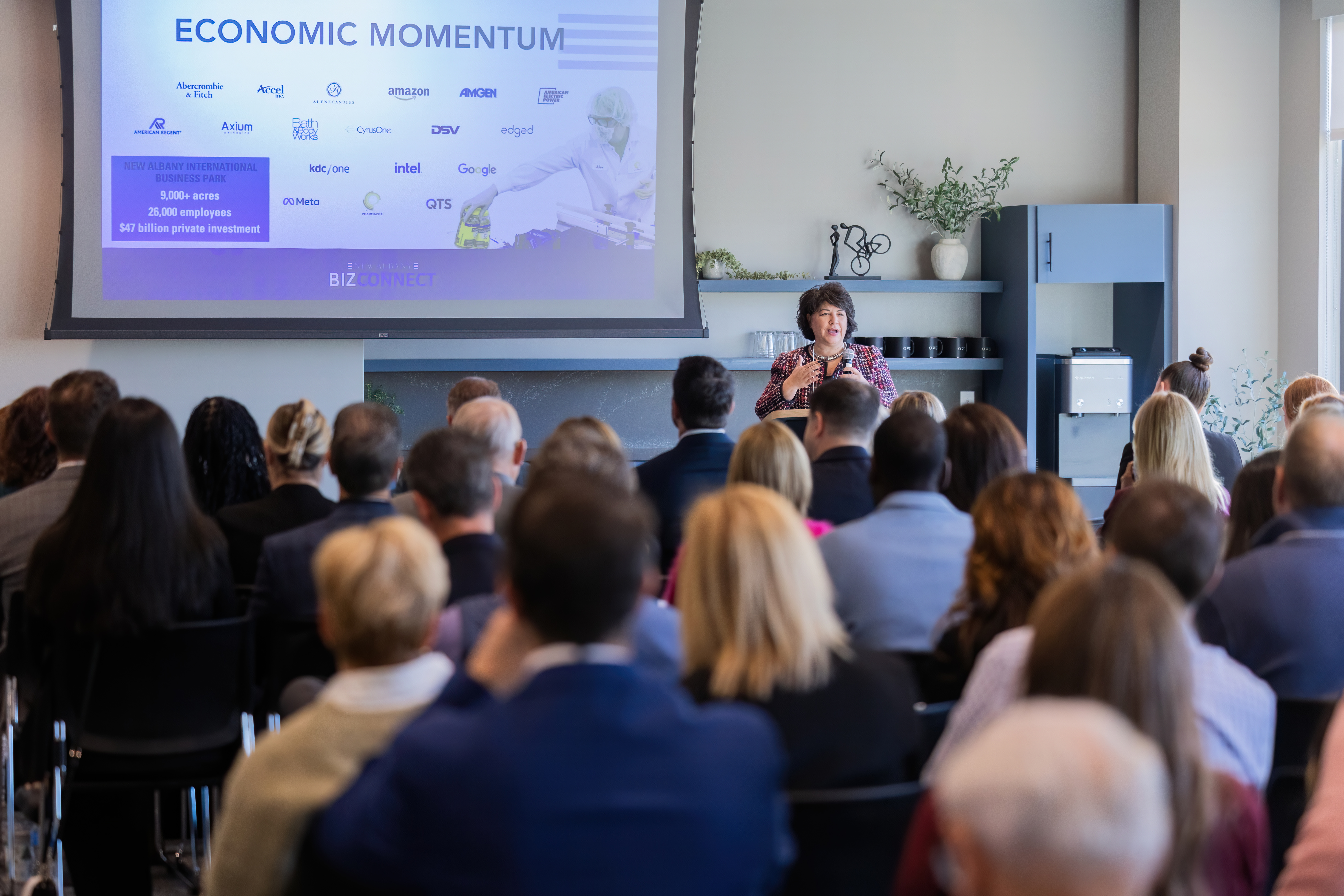 A presenter speaks in front of a crowd in a room. The speaker is standing in front of a slideshow