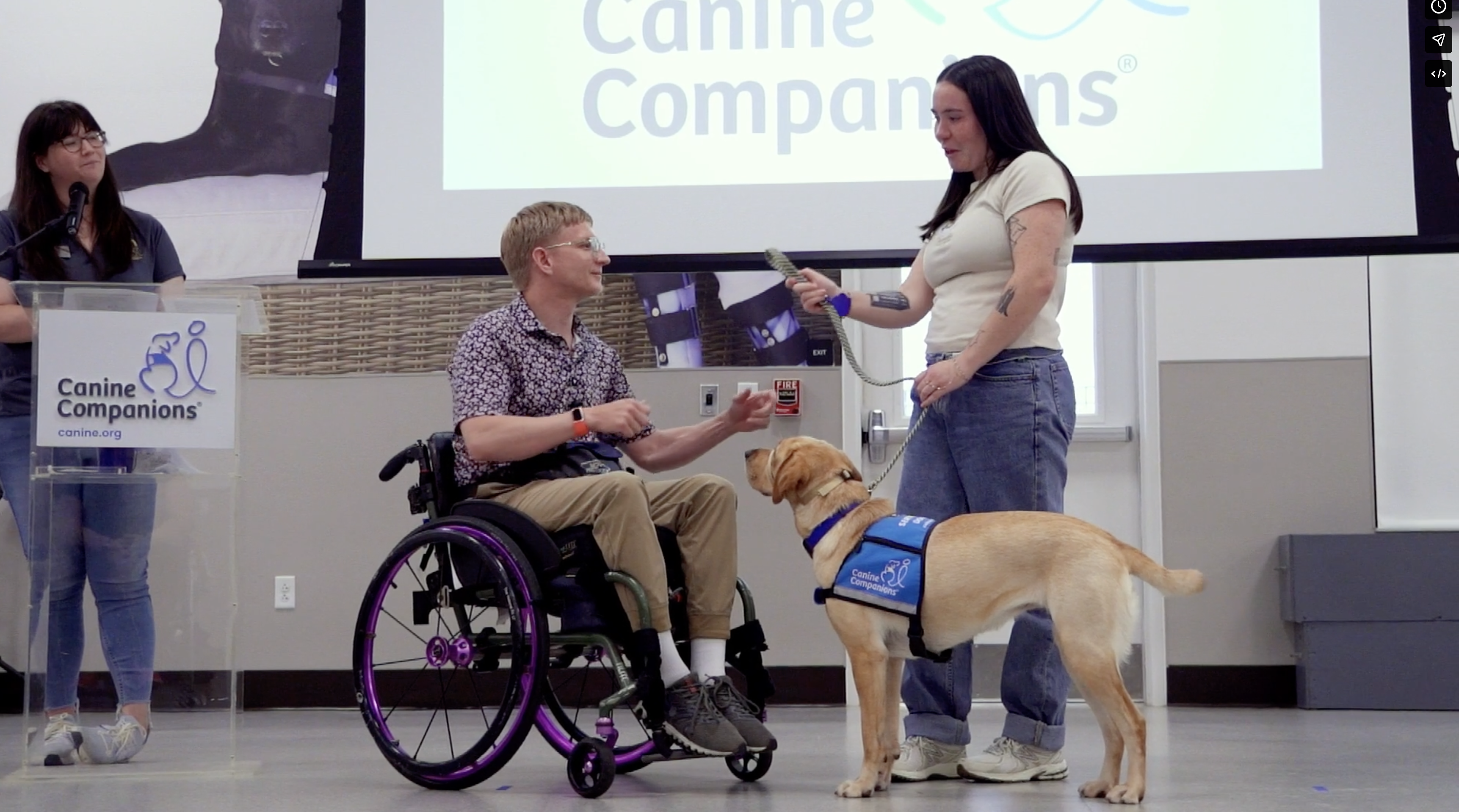 Conference presentation with a speaker standing beside a seated person in a wheelchair and a handler guiding a tan service dog wearing a blue harness